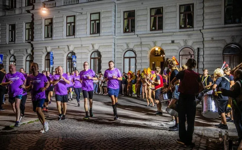 Midnattsloppet-löpare i lila springer förbi musiker och festklädda åskådare.